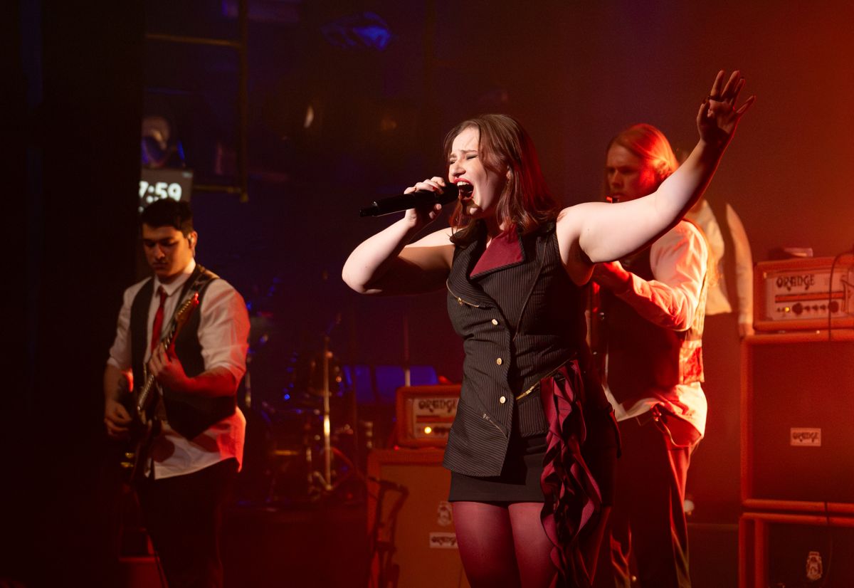 Fifi singing with her arm outstretched, the guitarist and amps behind her