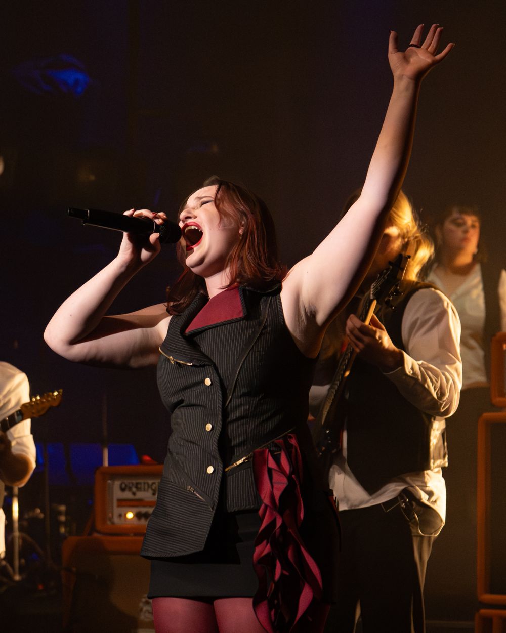 Fifi with her arm raised, singing in front of the guitarist and amplifiers